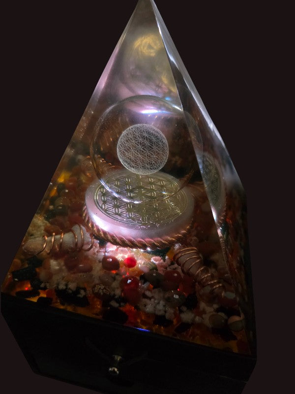 Close-up of Forge of Light orgonite pyramid lamp against a dark background, highlighting the etched Flower of Life glass sphere, selenite disc, copper detailing, and crystal inclusions softly illuminated.
