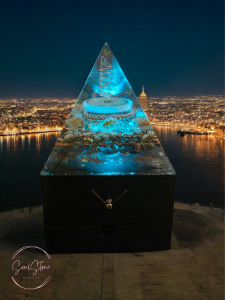 Blue-lit pyramid-shaped Lumen Drift Orgonite Lamp displayed at night overlooking a glowing cityscape; copper coil and crystals visible inside, sitting on a wooden jewellery box base with the SoulStone Designs logo