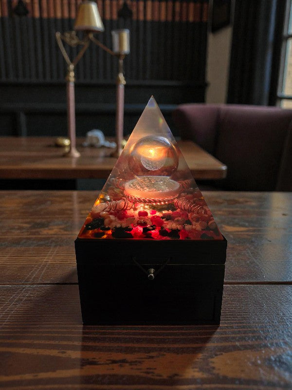 Forge of Light orgonite pyramid lamp glowing warmly indoors, featuring a glass sphere with 3D Flower of Life above a selenite disc, copper coil accents, and crystal layers on a black wooden base.