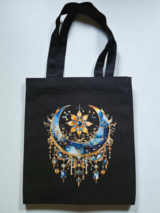 A black canvas tote bag with an intricate celestial design featuring a crescent moon, golden starburst, and gemstone-style hanging ornaments