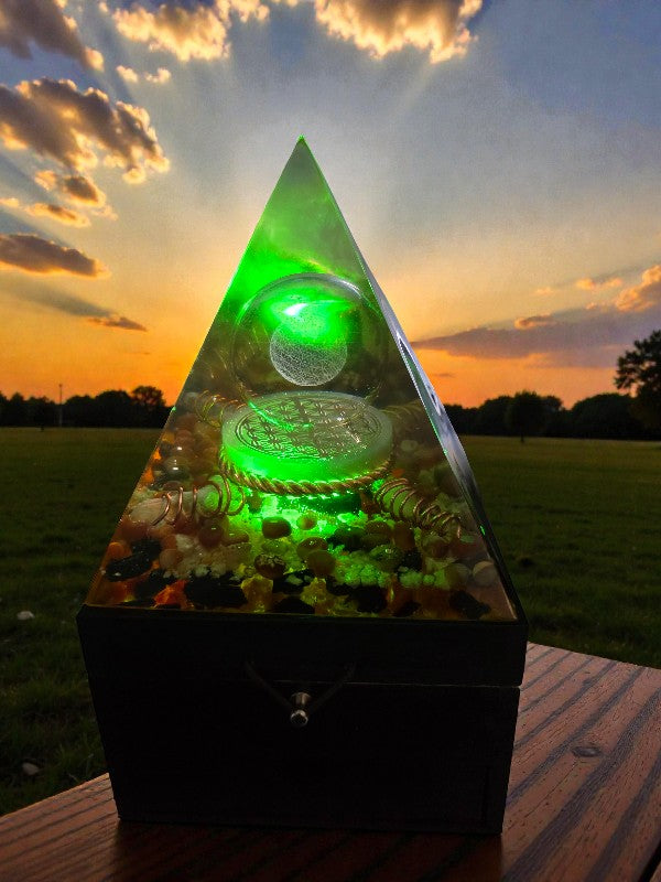 Forge of Light orgonite pyramid lamp illuminated green at sunset, showcasing a 3D Flower of Life glass sphere, selenite disc with copper Flower of Life, and copper-wrapped quartz points within a clear resin pyramid.