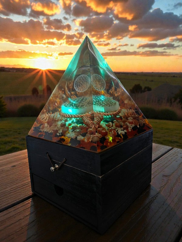 Forge of Light orgonite pyramid lamp glowing teal during golden hour, with a central Flower of Life glass sphere, copper tensor ring, selenite disc, and layered crystals inside a clear pyramid on a wooden surface.