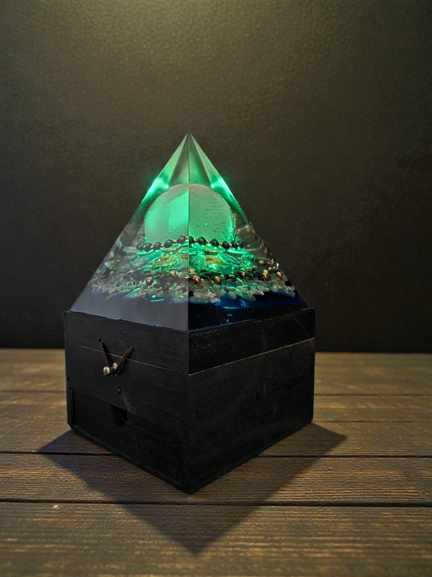 Axis of Stillness orgonite pyramid lamp illuminated green against a dark background, highlighting the central selenite sphere, hematite and copper bracelet, copper spiral, and crystal layers within clear resin.