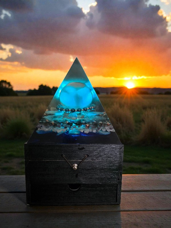 Axis of Stillness orgonite pyramid lamp glowing blue at sunset, showcasing a selenite sphere, grounding hematite accents, copper spiral, and soothing crystal inclusions on a wooden surface.