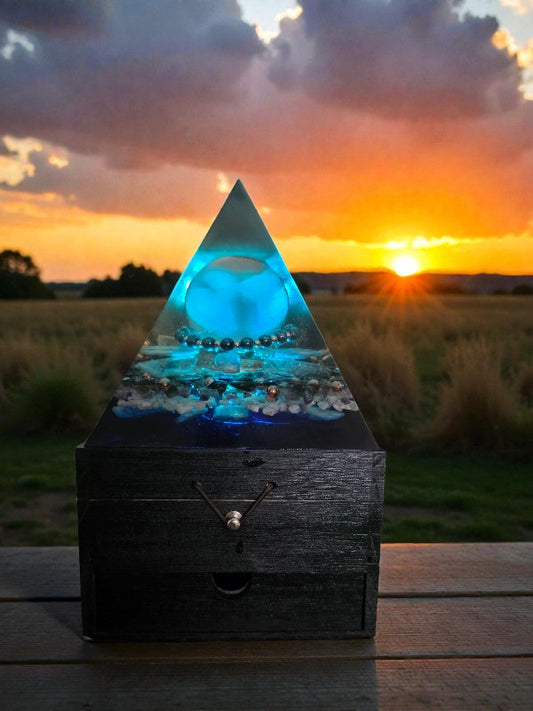 Axis of Stillness orgonite pyramid lamp glowing blue at sunset, showcasing a selenite sphere, grounding hematite accents, copper spiral, and soothing crystal inclusions on a wooden surface.