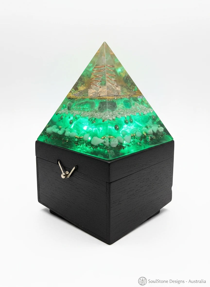 SoulStone Designs Celestial Clarity orgonite lamp with green crystals and copper spiral detail. Pyramid shape on a black wooden base.